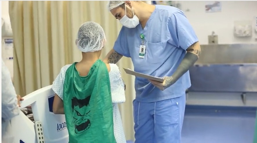 A imagem mostra um ambiente hospitalar com um médico com uniforme azul, touca e máscara de proteção; e uma criança, que está de costas, usando roupa hospitalar e uma capa verde com o rosto do incrível Hulk